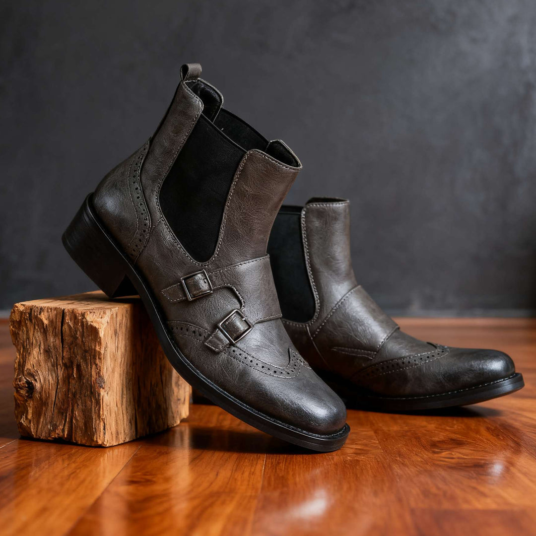 A pair of dark brown leather Chelsea boots with double buckle straps, displayed on a rustic wooden block over a polished wooden floor against a dark textured background.