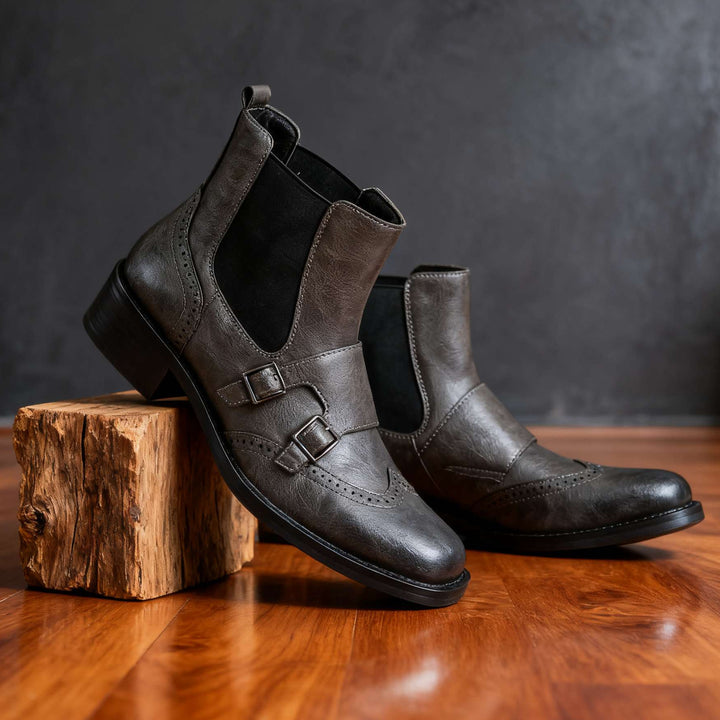 A pair of dark brown leather Chelsea boots with double buckle straps, displayed on a rustic wooden block over a polished wooden floor against a dark textured background.